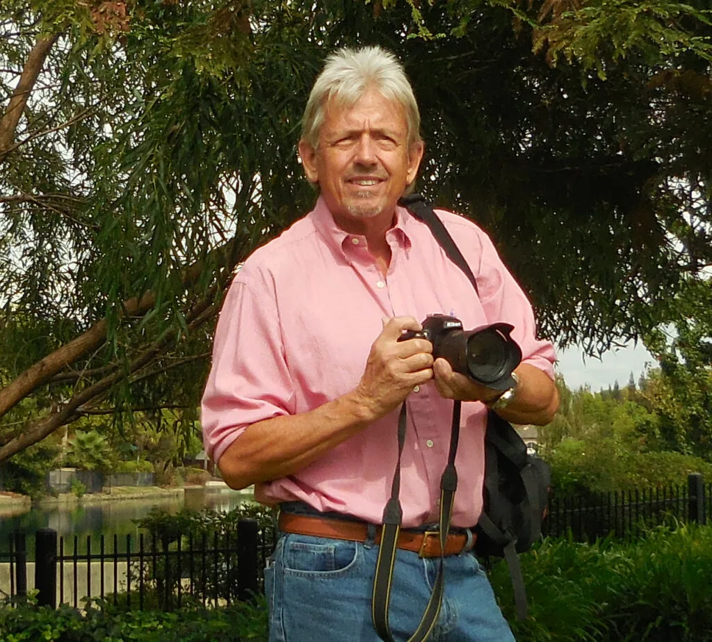Older man in pink shirt holding camera under willow tree by water