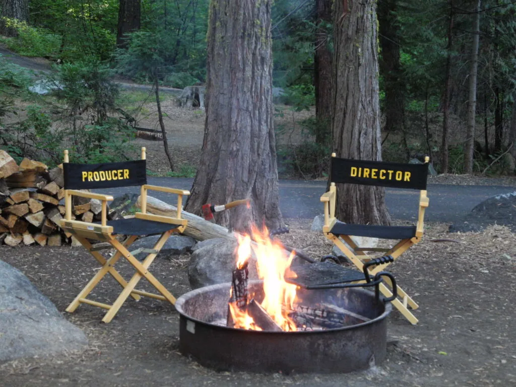 Two director's chairs labeled Producer and Director beside a campfire in a forest clearing with tall trees.