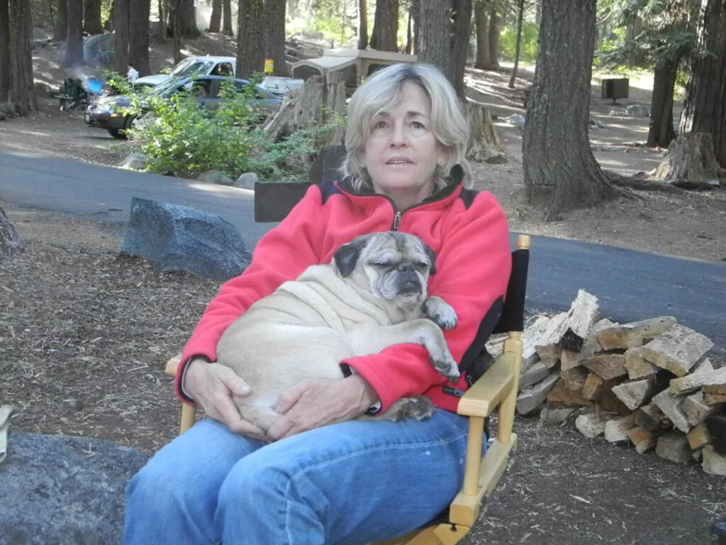 Woman in pink jacket sitting in camp chair holding small tan pug in forest setting