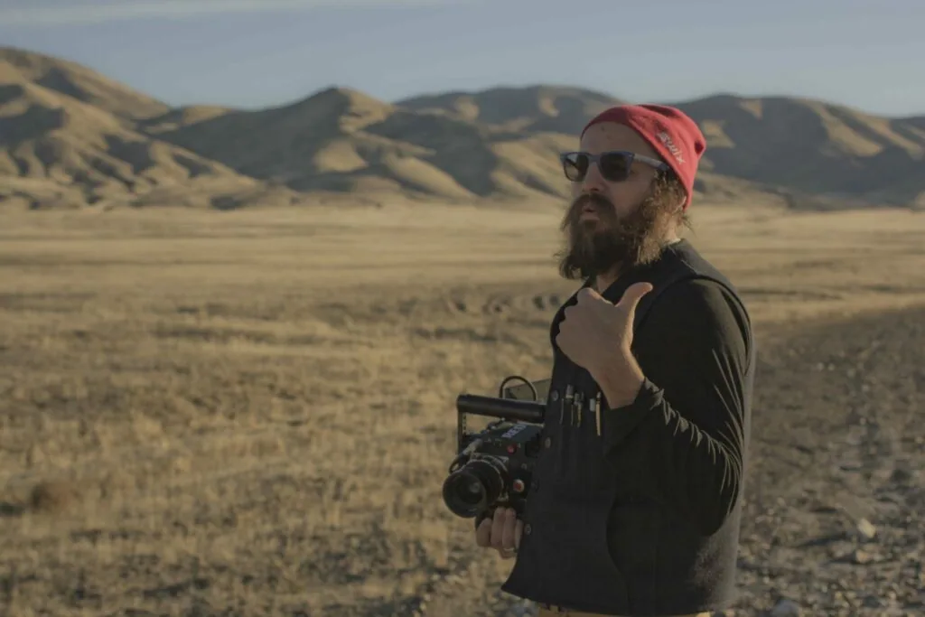 Bearded man wearing red cap and sunglasses holds film camera in arid desert landscape with mountains in background