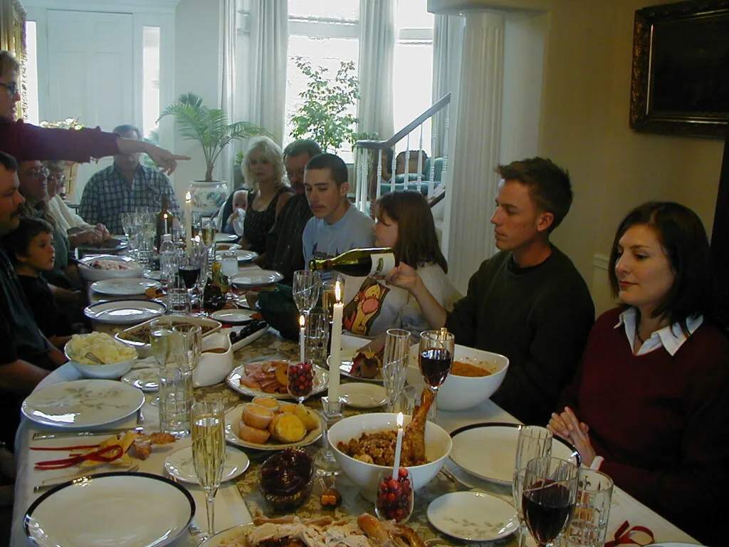 Group of people dining together at a long table set with plates, glasses, and food in a bright room with windows and curtains.