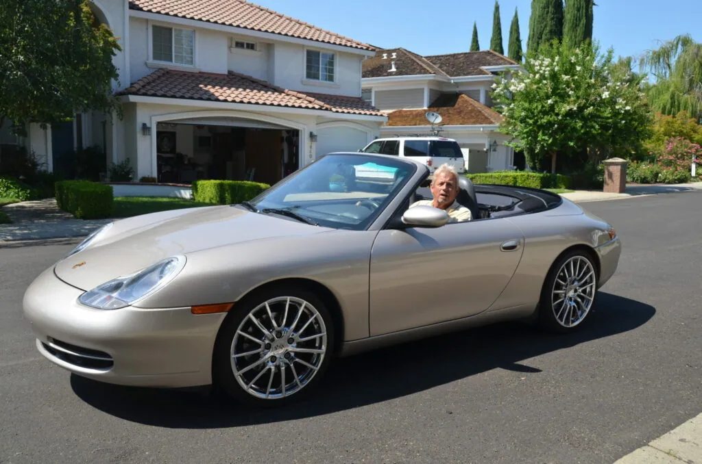 Silver convertible sports car parked in residential driveway with driver sitting in front seat, two-story white house with garage in background