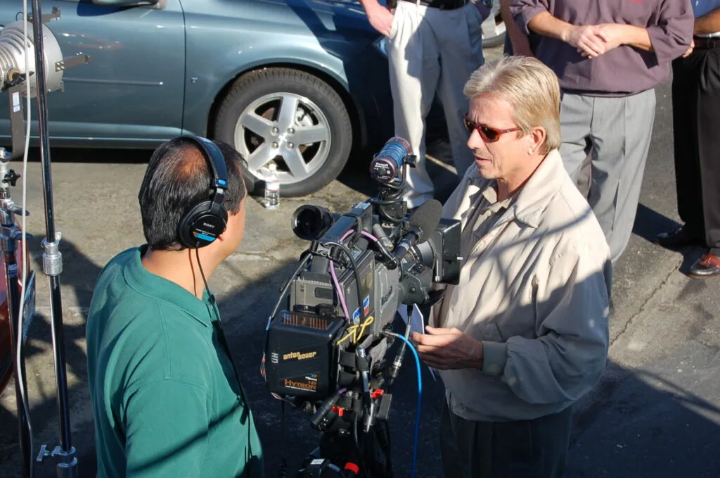 Man in sunglasses speaking into professional video camera microphone with operator in headphones at outdoor event