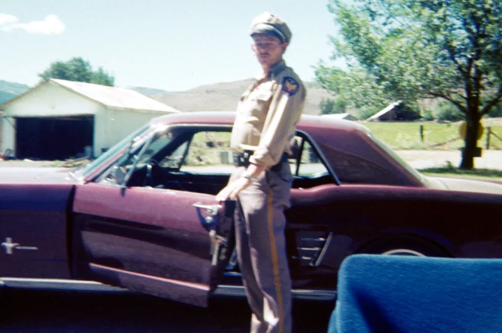 Officer in uniform standing beside a maroon car in rural setting with buildings and trees in background