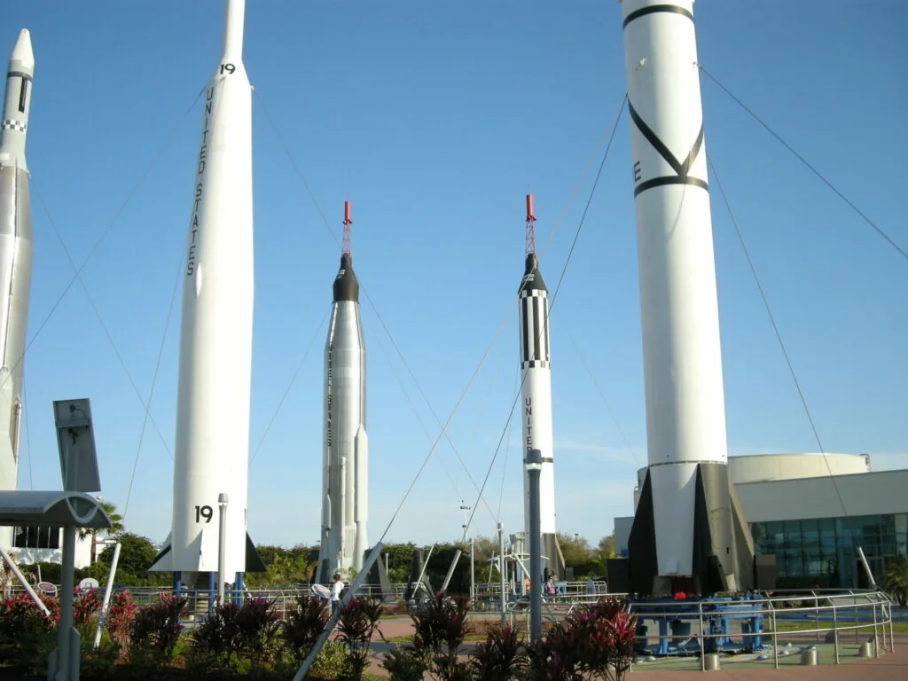 Collection of white rocket models displayed outdoors against clear blue sky, including tall cylindrical rockets with pointed nosecones and red tips