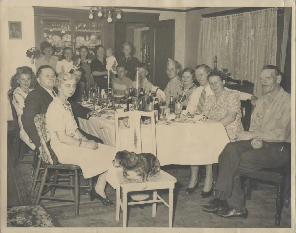 Group of men and women seated around a dining table with bottles and candles during a social gathering in a mid-century interior