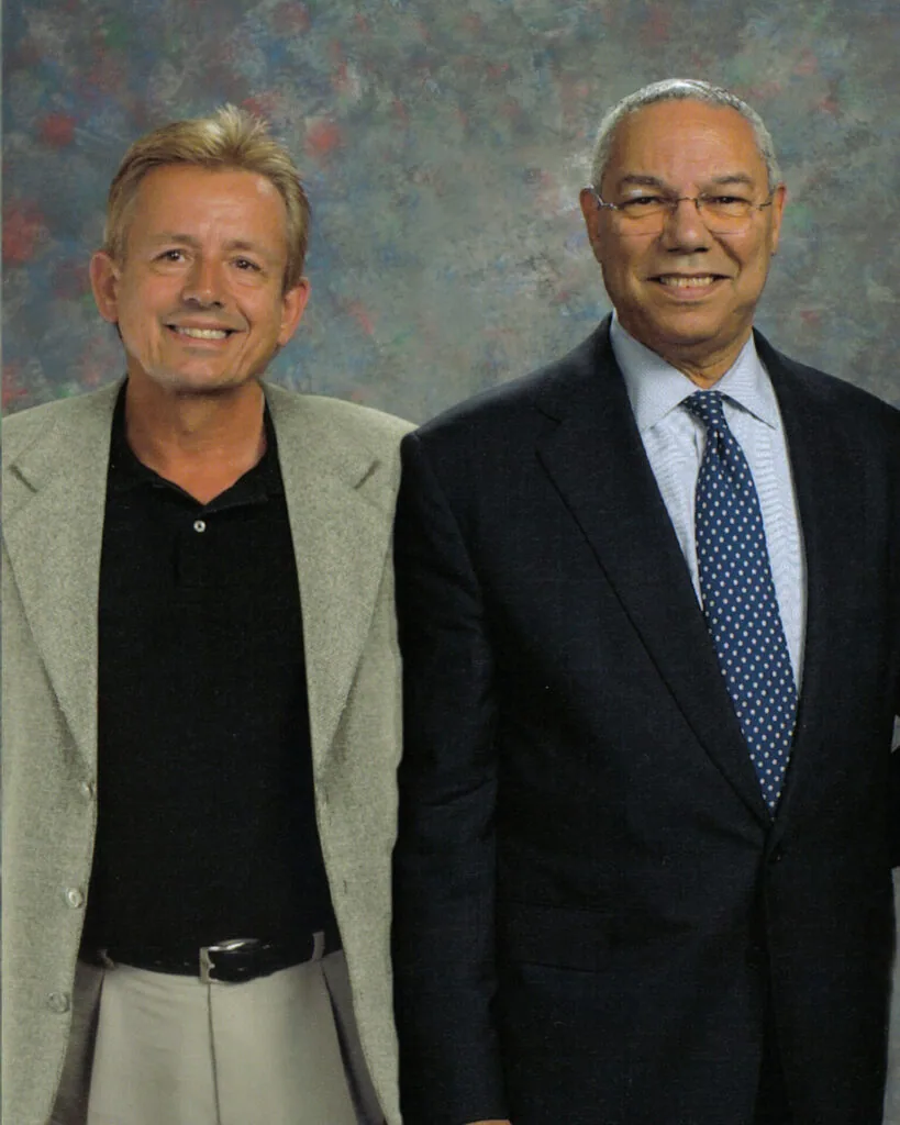 Two men posing together in front of a textured background, one wearing a beige blazer over dark shirt, the other in a dark suit with blue patterned tie