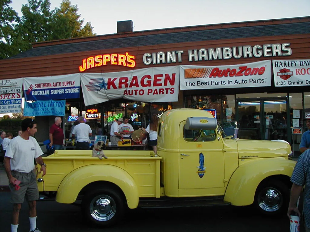 Vintage yellow pickup truck parked in front of Jasper's Giant Hamburgers restaurant with vintage neon signs and auto parts storefronts