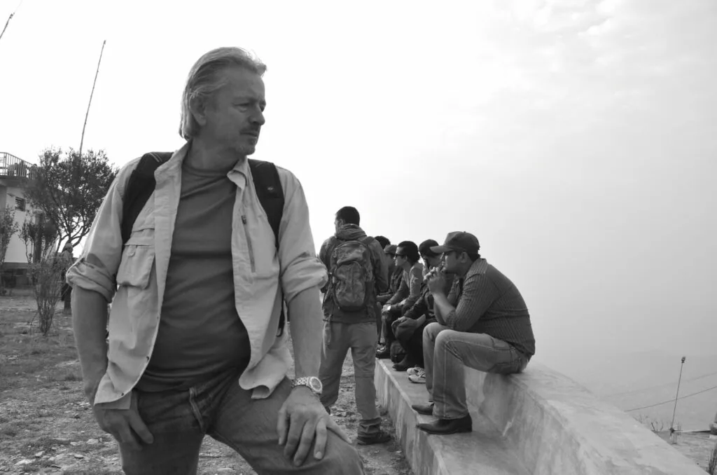 Man in light jacket crouching in foreground with group of people sitting on wall in background, black and white photograph