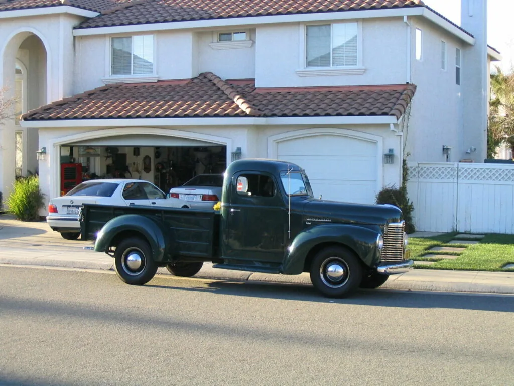 Vintage dark green pickup truck parked on residential street in front of two-story white house with clay tile roof