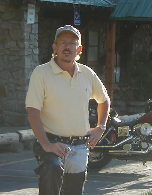 Man in yellow polo shirt and cap standing in front of motorcycle and stone building