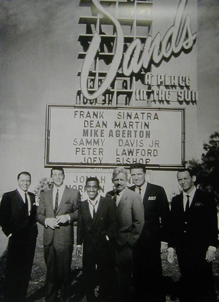 Six men in suits standing in front of a vintage movie theater marquee listing cast names including Frank Sinatra and Dean Martin.