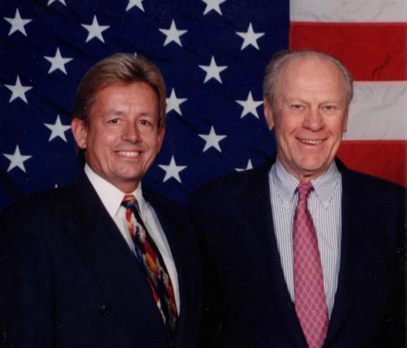 Two men in dark suits posing before an American flag backdrop