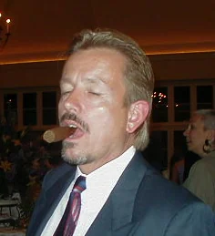 Man in dark suit and purple tie speaking at an indoor event with crowd in background