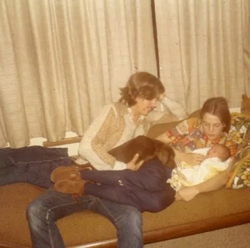 Two people sitting on a couch reading together, with a teddy bear visible in the foreground, beige curtains in the background