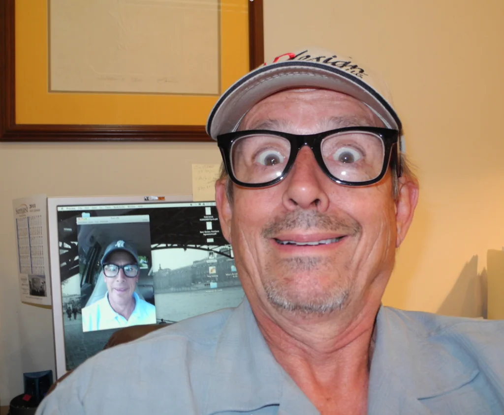 Man wearing glasses and cap smiling at camera with computer monitor showing video call in background