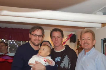 Four men posing together indoors, one holding a doll, with holiday decorations visible in the background