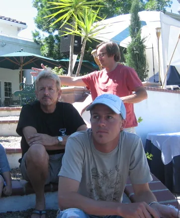 Three people gathered outdoors in a residential setting with palm trees and white structures visible in the background.