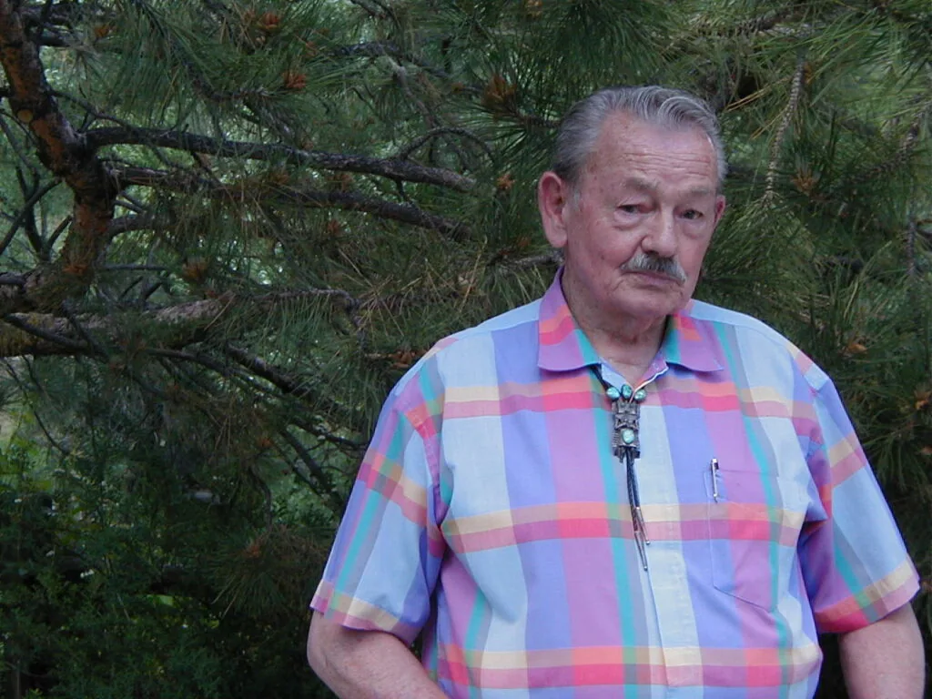 Older man with gray hair and mustache wearing multicolored plaid shirt with bolo tie, standing in front of evergreen trees