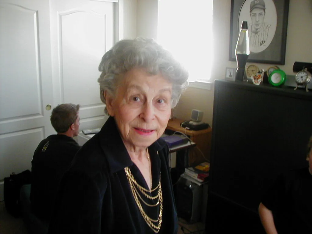 Elderly woman with gray hair wearing black jacket and gold chain necklaces, smiling at camera in home interior