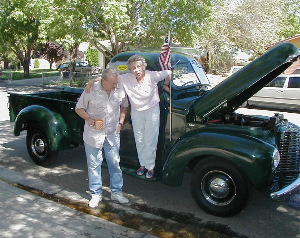 Two elderly people standing beside a restored green vintage truck with hood open at an outdoor car show, American flag visible on vehicle