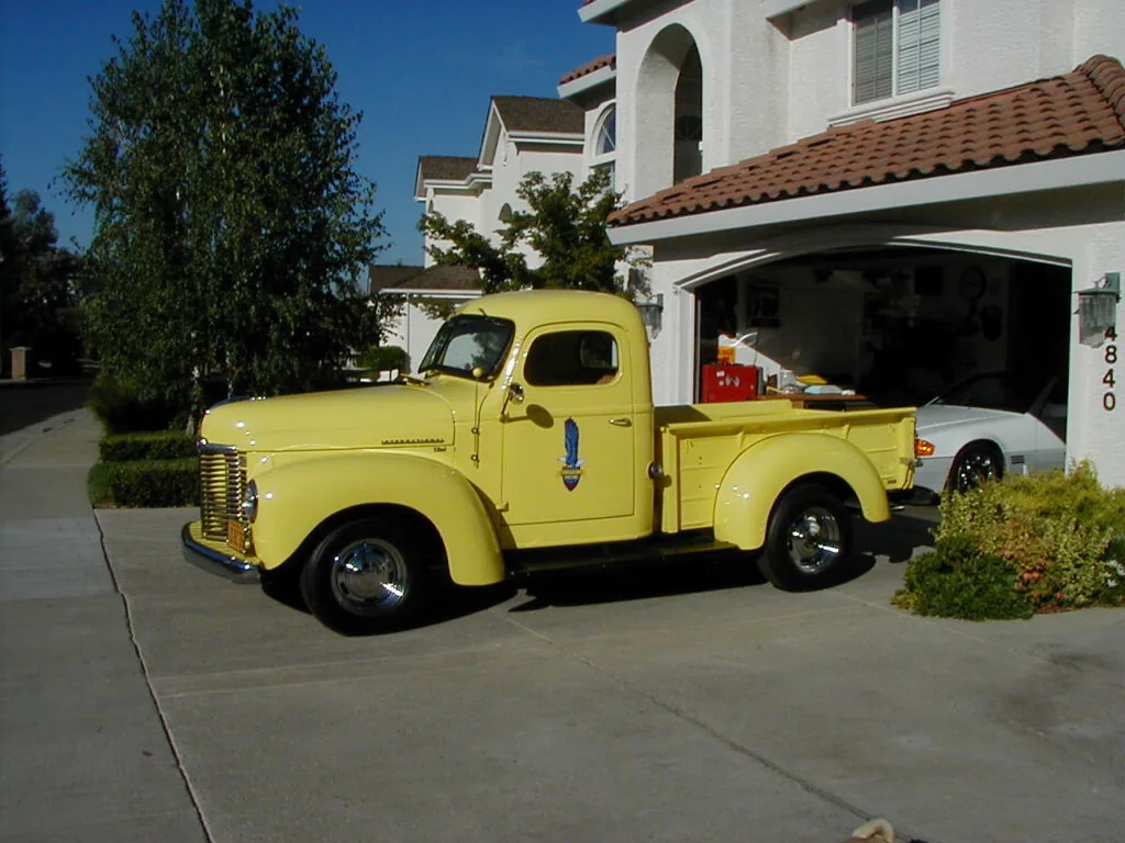 Bright yellow vintage pickup truck parked in driveway beside white house with red tile roof