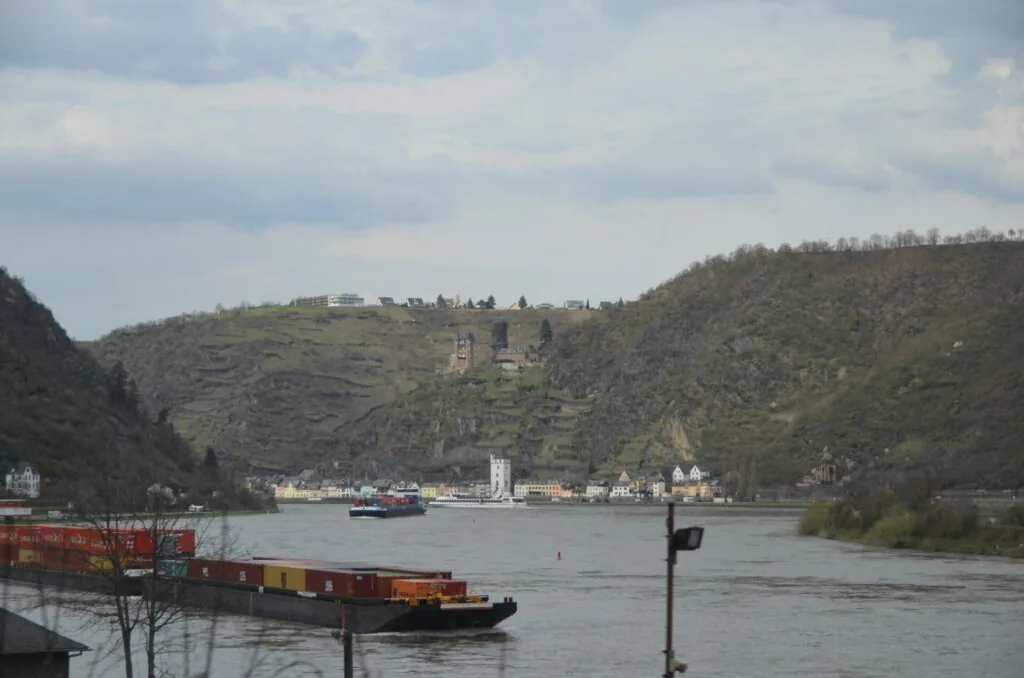 Cargo barge on river with forested hillside, castle ruins, and riverside town buildings under cloudy sky