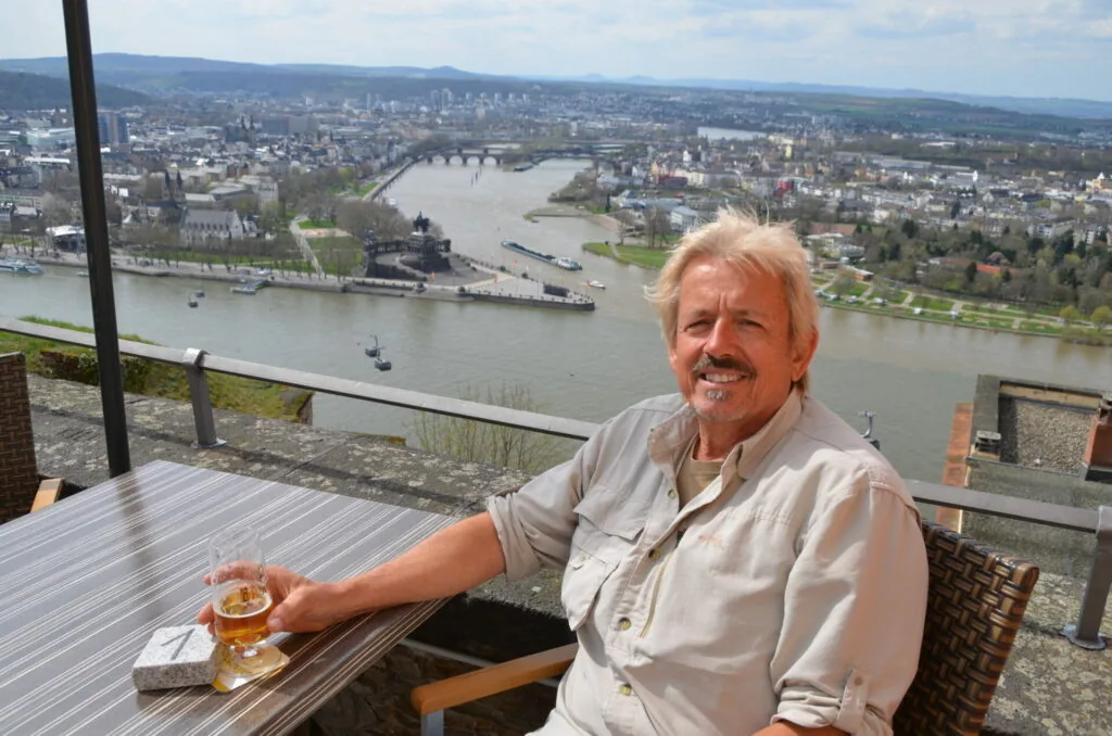 Man holding a drink at an outdoor table overlooking a river valley with a city in the distance