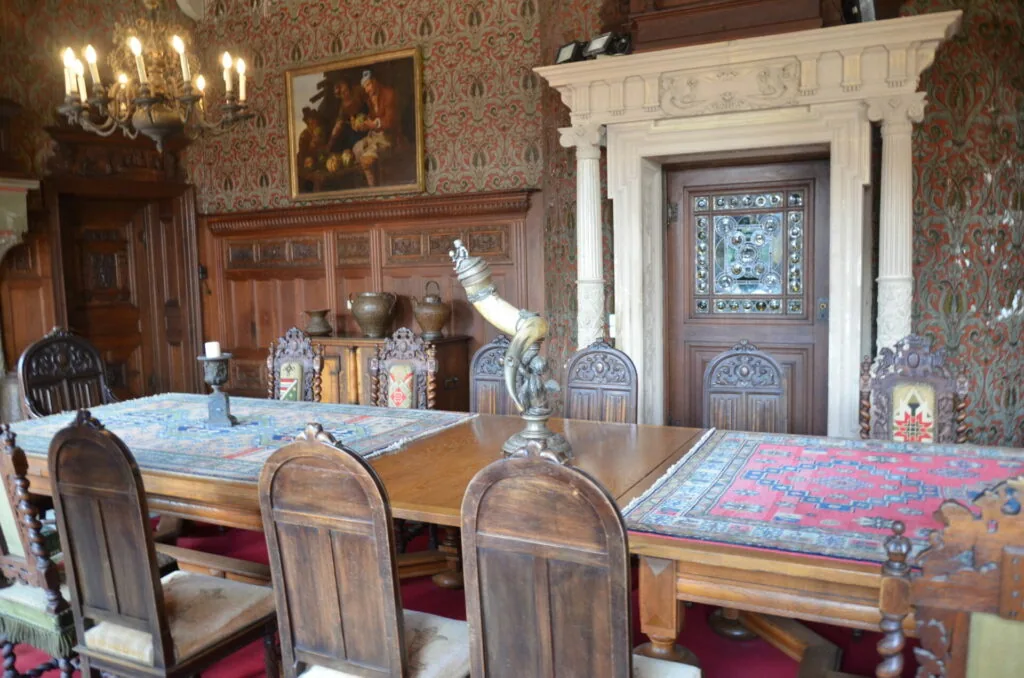 Historic dining room with ornate wallpaper, wooden furniture, chandelier, fireplace mantel, and decorative objects on display shelves