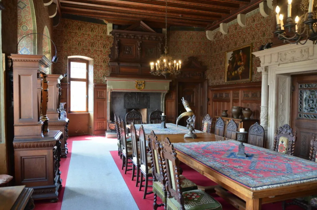 Historic dining room with wooden beams, patterned wallpaper, chandelier, long table with patterned runner, wooden chairs, fireplace, and period furnishings.