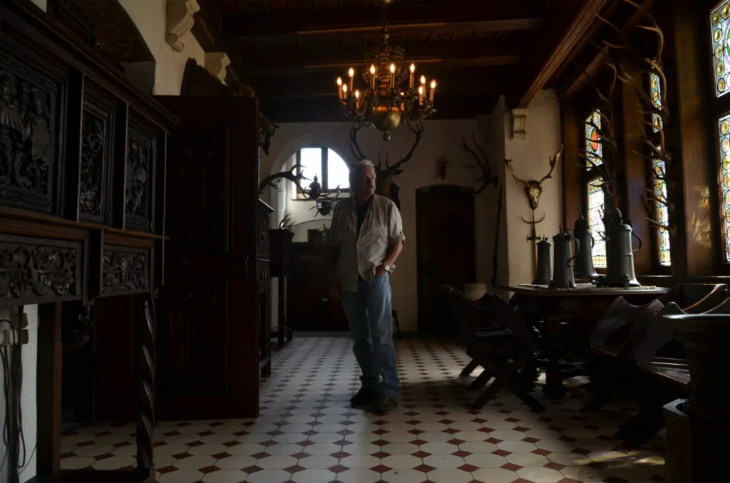 Man standing in ornate room with dark wood cabinets, mounted antlers, stained glass windows, and chandelier