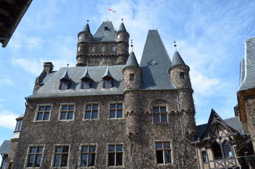 Historic stone castle with multiple towers, turrets, and slate roofs against blue sky with clouds