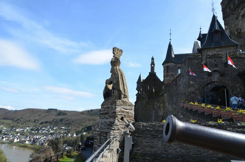 Stone statue and cannon on castle ramparts overlooking river valley with medieval castle towers and flags visible