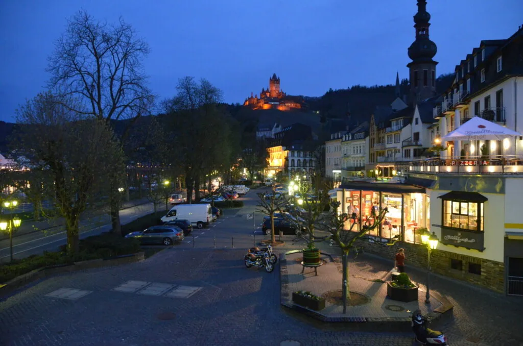 Twilight street scene with illuminated storefronts and castle on hilltop in background