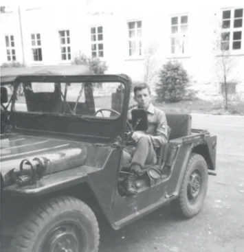Man sitting in open-top military jeep parked in front of brick building with windows