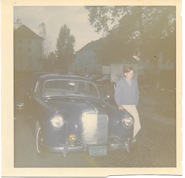 Man standing next to dark sedan automobile in front of residential buildings and trees