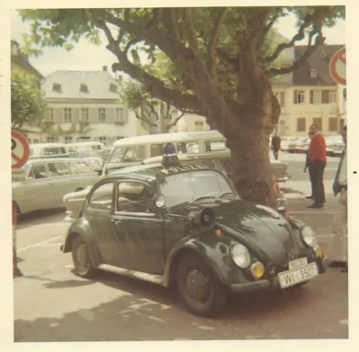 Dark Volkswagen Beetle parked under a large tree on a European town square with period cars and buildings visible in background