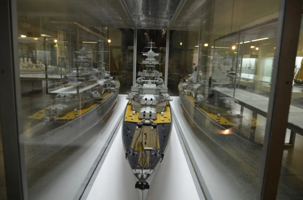 Model military warship with yellow and gray hull displayed in museum glass case with other ship models visible on sides