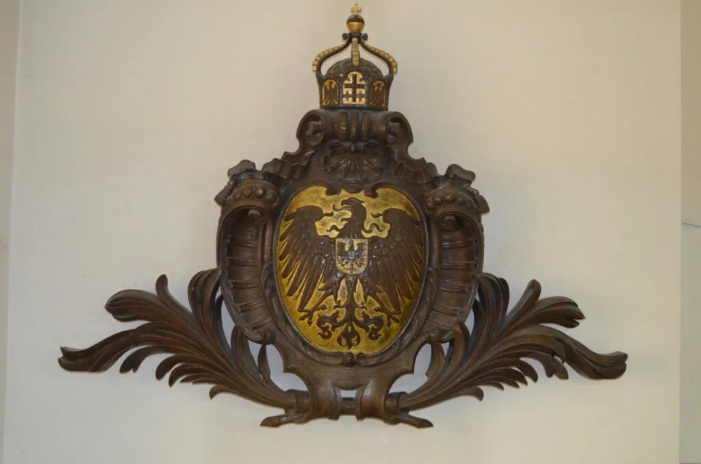 Ornate wooden coat of arms with golden eagle emblem, crown top, and decorative scrollwork flanked by carved lions