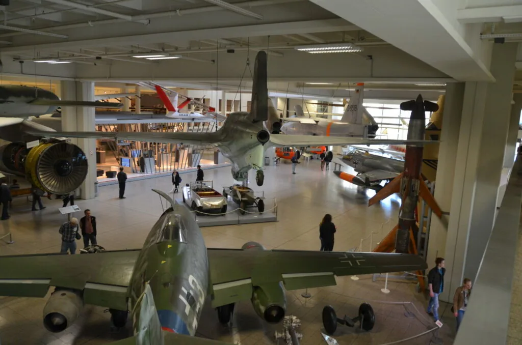 Interior of aviation museum with military aircraft on display, including planes with propellers and visitors walking through the hangar space
