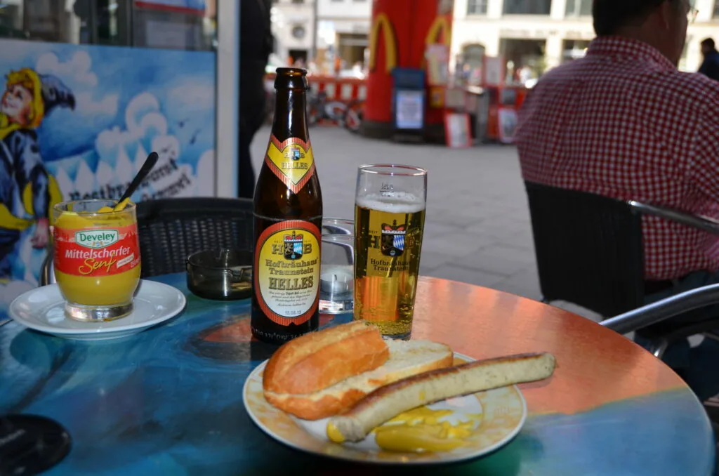 Beer bottle and glass with sausage, bread roll, and mustard on outdoor table at street festival