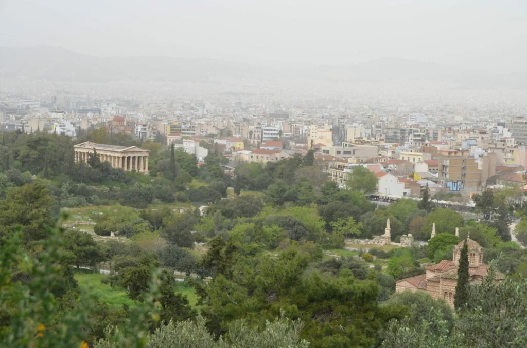 Panoramic cityscape with hazy skyline, green vegetation in foreground, classical temple structure on left, and dense urban buildings extending to horizon