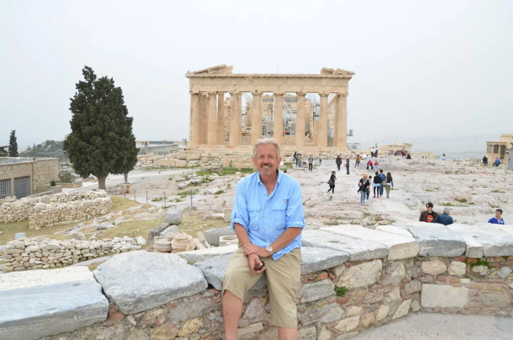 Man in light blue shirt sitting on stone wall with ancient Greek temple ruins in background