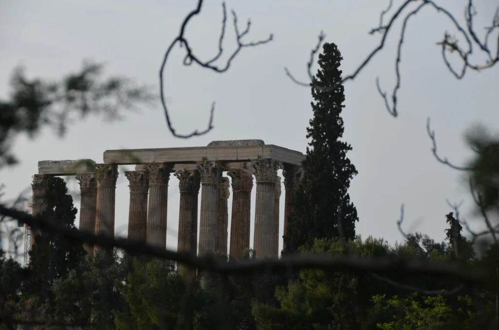 Ancient Greek temple ruins with standing columns framed by bare branches and cypress trees against overcast sky