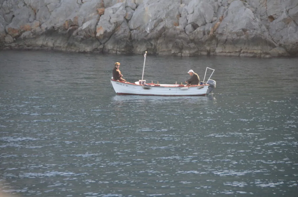 Two people in a white fishing boat on calm water near rocky cliffs
