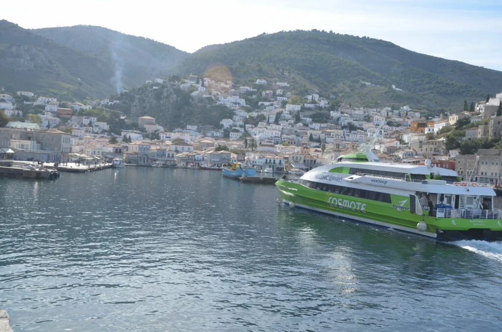 Green and white ferry boat docked in Mediterranean harbor with white hillside buildings and forested mountains behind