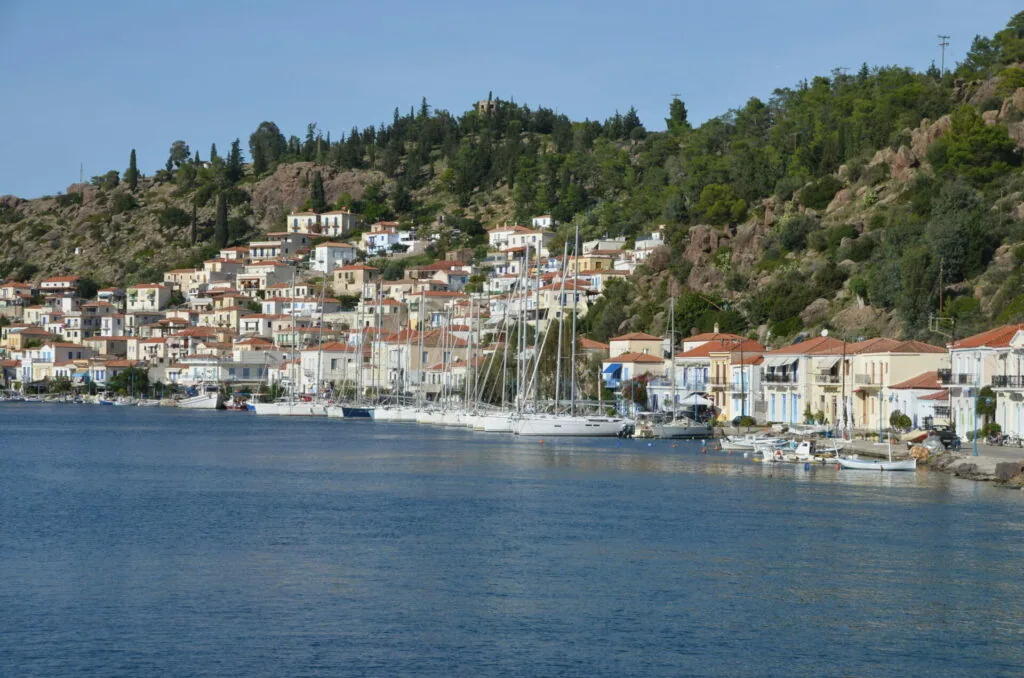 Waterfront village with colorful multi-story buildings and sailboats moored along a blue harbor, backed by forested hills.
