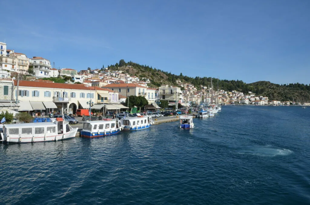 Waterfront harbor with fishing boats, waterfront buildings, and hillside homes with coniferous trees under clear blue sky