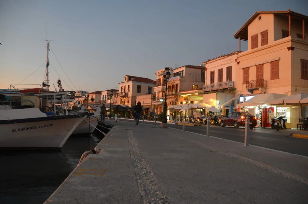 Mediterranean harbor at dusk with moored fishing boat, terracotta buildings, and waterfront shops with warm lighting