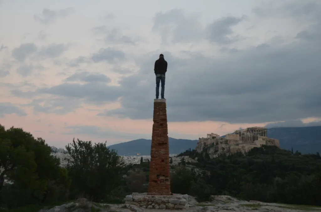 Person standing atop a tall brick column overlooking a city with mountains and an ancient acropolis in the background under a cloudy sky